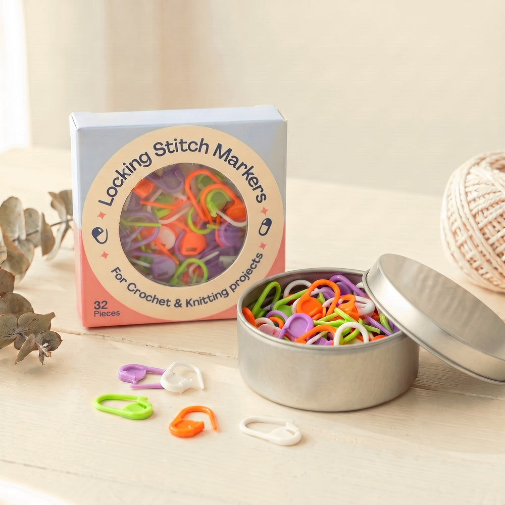 Crochetree locking stitch markers box standing next to an open silver tin of markers, with loose markers on a cream table